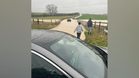Cars in floodwater on a rural road. Some people on foot are in attendance.