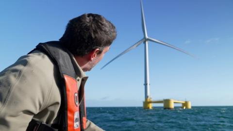 Climate editor Justin Rowlatt in foreground looking at a floating turbine in the background