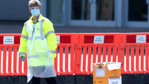 CARDIFF, WALES - APRIL 07: A worker wearing a surgical mask and apron stands near a test kit drop-off area at a drive-through testing centre at the Cardiff City stadium on April 7, 2020, in Cardiff, Wales. The testing centre opened today for frontline staff and people in the Gwent area. There have been over 50,000 reported cases of the COVID-19 coronavirus in the United Kingdom and over 5,000 deaths. The country is in its third week of lockdown measures aimed at slowing the spread of the virus.