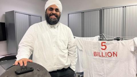 Man with beard wearing turban and standing next to washing machine and t-shirt which states "5 billion"