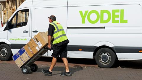 A Yodel parcel delivery driver walks in front of a white Yodel van with parcels in a reflective vest in 2018