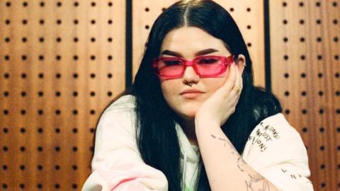 Woman with long dark hair and wearing white top with red glasses sits with her chin in her palm