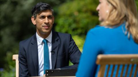 Rishi Sunak in an interview with the 91ȱ's Laura Kuenssberg. He is sitting outside in a suit across form a garden table, with the back of the host visible in the foreground, with her face and body blurred.