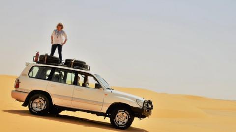Sue Rogers stands on the top of a car in the dunes of Sudan