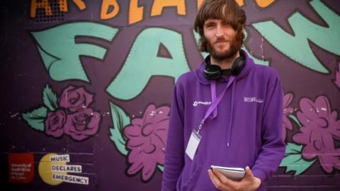 Johnny Giles in a hoodie with long bangs and a beard standing in front of a mural, holding a note pad