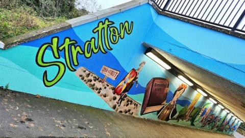 An underpass with a mural on one side, with a background in different shades of blue with paintings of a Roman soldier, the Domesday book and a church