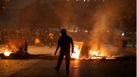 A riot police officer walks near burning fire during protests in Beirut