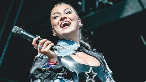 Caity Baser performing on stage. Caity is a 21-year-old woman, she has her long blonde hair tired back and wears a denim shirt over a cropped black latex top with a silver star on it. She has lots of earrings and wears pink lipstick and graphic green eyeliner and leans her head back and smiles at her audience while holding her microphone away from her with her right hand.