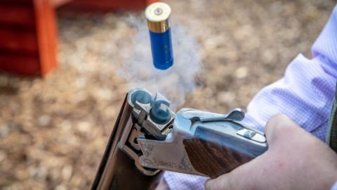 A blue shotgun cartridge jumps into the air, ejected from the smoking barrel of a shotgun broken open on its hinge to reload