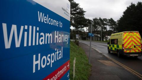 An ambulance driving by a sign for the William Harvey Hospital in Ashford, Kent