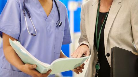 A nurse in a blue uniform and a hospital worker in a smart jacket look together at an open folder