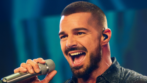 A man with close-cropped brown hair, beard and brown eyes sings into a microphone. He has a black earpiece in his left ear. He is wearing a sparkly black shirt.