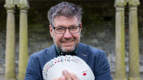 Dr Gustav Khan who has a brown and grey beard, gelled black and grey hair and is wearing black-rimmed square glasses displays a pack of cards towards the camera with a slight grin on his face.