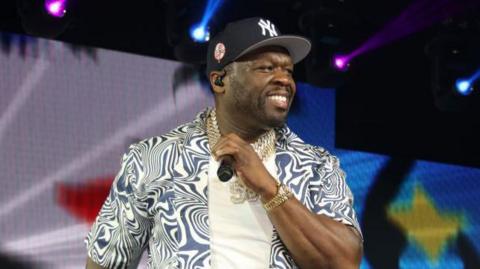 Rapper 50 Cent looks off stage while holding a microphone. He is wearing a blue and white patterned shirt, a white t-shirt and gold jewellery