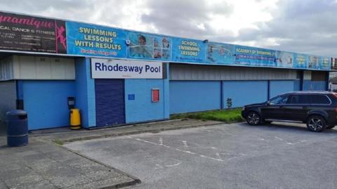 A single storey blue building with parking spaces in front and adverts for swimming and fitness lesson on attached hoardings