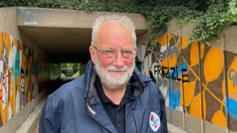 A man looking at the camera. He has a white beard and white hair. He's wearing glasses and a navy jacket. Behind him are murals on the walls of a tunnel under the road, covered in graffiti.