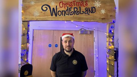 Jason smiles at the camera while standing below a wooden arch with a handmade sign saying "Christmas Wonderland". He wears a red and white Santa hat and a black airtex T-shirt with the academy's golden logo on. Fairy lights wind up the archway.