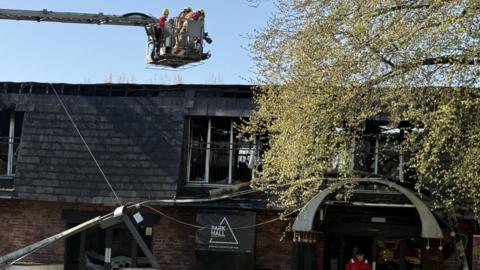 A number of firefighters are suspended above a fire-damaged building in a cherry picker.