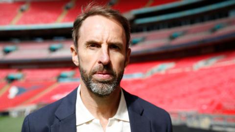Gareth Southgate, wearing a dark blazer and white shirt, standing in an empty football stadium with red seats