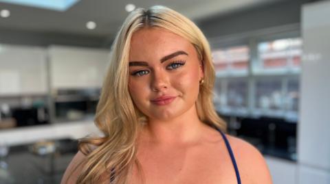 The young women has long, blonde hair falling over her right shoulder. She is wearing a blue dress with thin straps and gold hoop earrings. She has tanned skin and blue eyes and is looking straight into the camera with a closed-mouth smile.