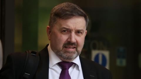 Robin Swann, who has short dark hair and beard, leaves a building wearing a dark suit jacket, a white collared shirt and a purple tie. He is carrying a black backpack strap on his right shoulder.