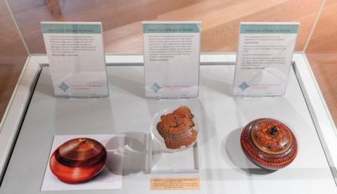 A display case containing three objects with information cards. On the left, there is a photograph of a round, striped container with earthy tones. In the middle, there is a fragment of an ornate, reddish-brown artefact and on the right, there is a brightly coloured, circular container with intricate red, black, and orange patterns and a small knob on top.