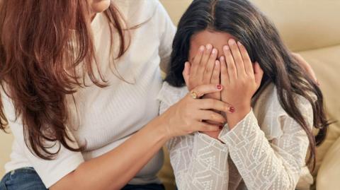 A girl with long dark hair is covering her face with her hands. A mother is comforting her with one hand resting on the girl's hand. Her auburn hair is partially obscuring her face. They are both sitting on a cream coloured sofa. 