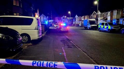 A street photograph of St Saviours Road. It is dark outside and police cordon tape can be seen across the road, with a police van and car parked in the cordon 