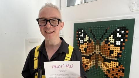 A man, Clarke Reynolds, holding a piece of paper reading "Touch Me!" in front of a butterfly art design made up of lots of dots.