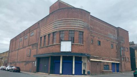 A large red‑brick corner building on a quiet street under a grey, overcast sky. The structure has a curved façade at the corner, with horizontal decorative bands and tall windows on the upper level.
On the ground floor, several blue metal roller shutters line the building, suggesting former commercial or storage use. A few cars are parked along the street to the left, and the roads around the building are empty.