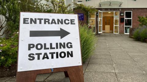 A picture of a wooden sign which reads 'Entrance' with an arrow pointing to the right, with the words 'Polling Station' below.