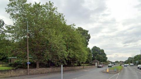 Bradford Road in Cleckheaton. Pictured is some trees on the left-hand side of the image, with a main road to the right. The carriageway ahead is divided by a grassed traffic island