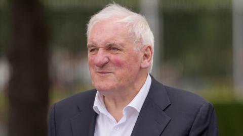 Bertie Ahern, a man with short, white hair, looks past the camera into the distance during a 2025 event. He is wearing a a navy suit jacket and a white shirt. The background is blurred