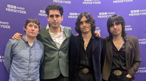 A group shot of Los Mapaches. Jevan Turner is on the left. He has short dark hair and is wearing a denim shirt. Jose Rozas has short dark curly hair and is wearing a green jacket. Fabrizio Rozas has long curly hair and is wearing an open shirt and a dark jacket. Piero Rozas has long dark hair and is wearing an open shirt and brown jacket.