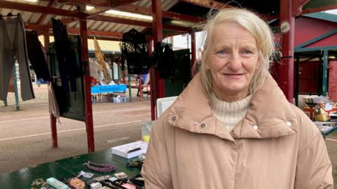 A woman in a faded pink coat standing next to a a market stall. 