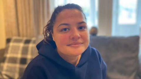 Vicky Prussia, wearing a blue hooded top, looking at the camera while sitting in a living room