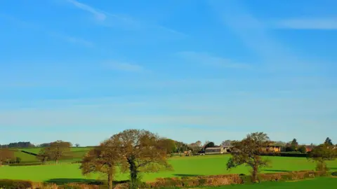 Blue skies and sunshine over a countryside scene with fields, some buildings and trees in the foreground