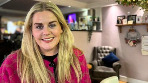 A woman with long blonde hair and a pink coat is sitting in a cafe area. She is smiling. Behind her there are chairs and tables and some greenery. There are also sleeves of pictures and framed photographs. 