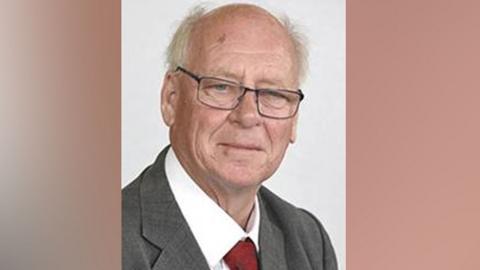 Head and shoulders shot of Chris Snow. He has short white hir, dark-framed glasses and is wearing a grey suit jacket, white shirt and dark red tie
