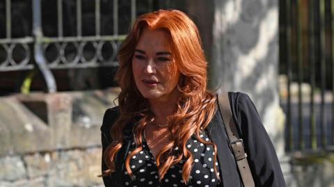 Anne Donaghy outside court. She has long red hair, wearing a necklace, black polka dot blouse and black blazer.