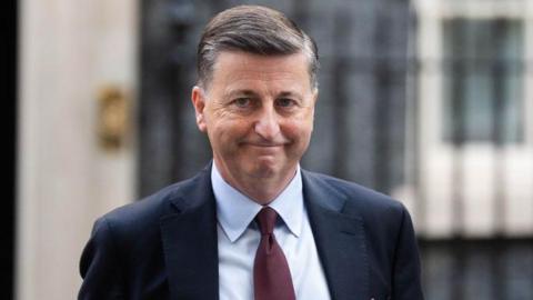 Douglas Alexander, who has short black hair, smiles as he looks towards the camera. He is wearing a dark suit, pale blue shirt and red tie, with a red folder tucked underneath his left arm. 