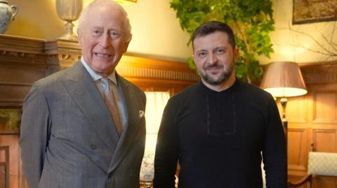 The King, stood on the left hand side, shakes hands with Zelensky in front of a fireplace in the Saloon of Sandringham House in Norfolk.