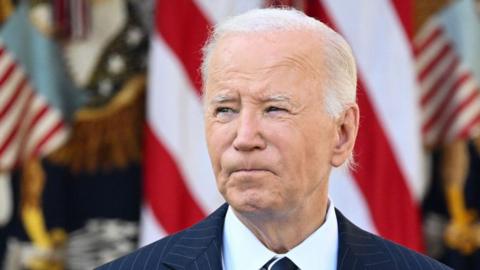 US President Joe Biden looks into the distance. He wears a pinstriped suit and has American flags behind him.