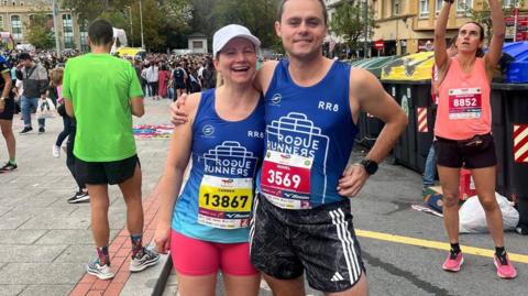 A woman and a man in running gear. There is a runner behind them taking a photo and there is a crowd behind them. 