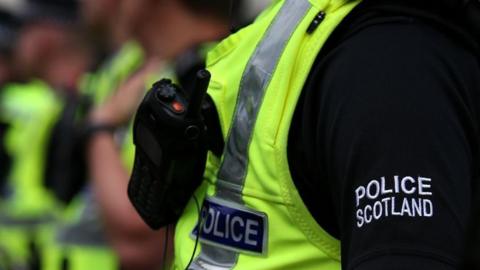 Generic image - Close up of a police officer wearing a black shirt with "Police Scotland" in white stitching on the sleeve and a high visibility vest with "Police" labelling on it and a walky talky strapped to their chest. In the background, other officers are visible but out of focus.