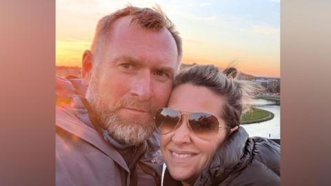 Justine, who is wearing sunglasses, is posing for a picture with her husband as the sun sets behind them. 