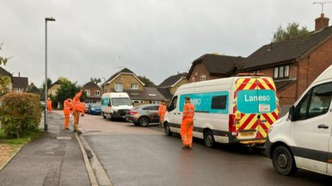 Vans line right hand side of residential street with workers in orange on pavement and in road with homes seen in background