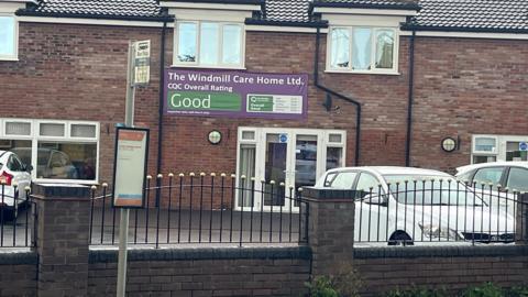 The outside of a bricked building, with cars parked outside, and a fence and brick wall in the foreground. There is a purple and green-coloured banner pinned to the building reading THE WINDMILL CARE HOME LTD.
