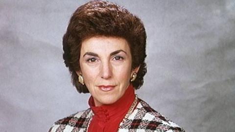 Woman wearing red polo neck shirt, chequered jacket and gold earrings stares directly.