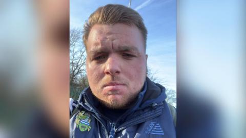 Jake Warner looks at the camera in a selfie. He is wearing a blue jacket with the Chertsey Town club badge on it and has his blond hair brushed back. The sky behind him is blue.
